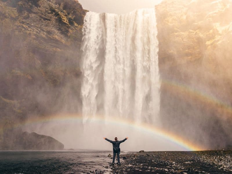 Person with their hands in the air as they admire the waterfall and rainbow in front of them.