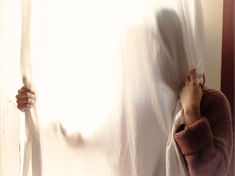 Woman hiding behind curtain representing feeling safer hiding from trauma than accepting it.