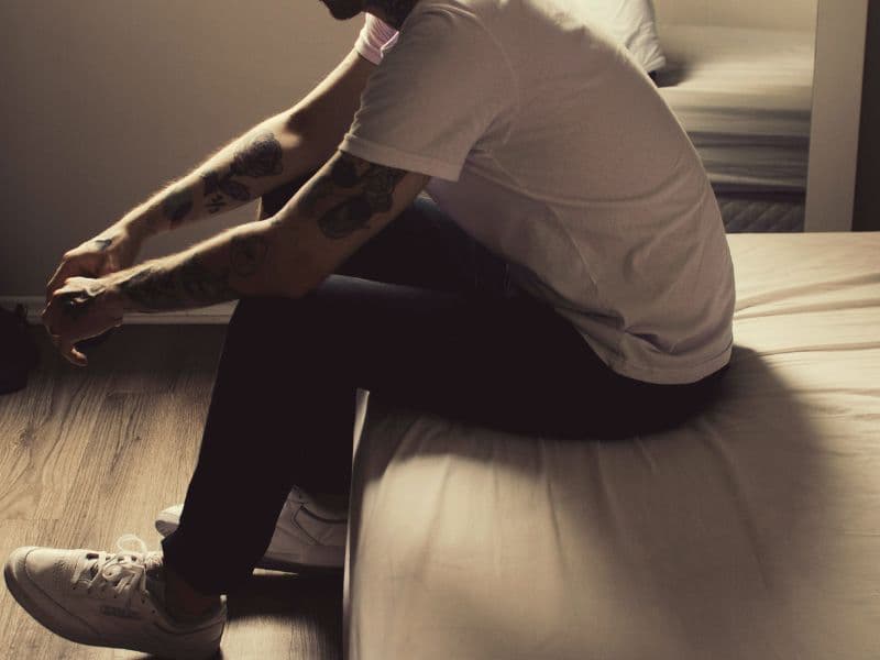 Man sitting on a bed with his hands placed by his knees appearing to be in deep thought.