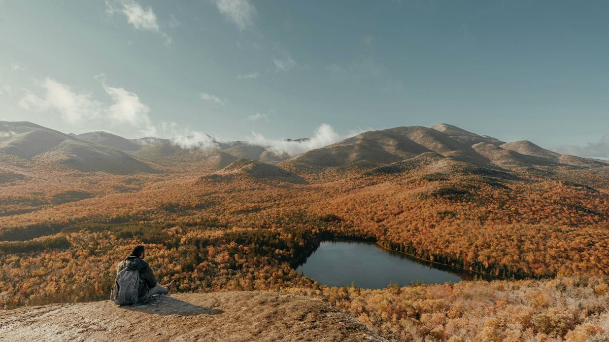 Person sitting quietly on a cliff overlooking the autumn forest and a pond, reflecting alone.
