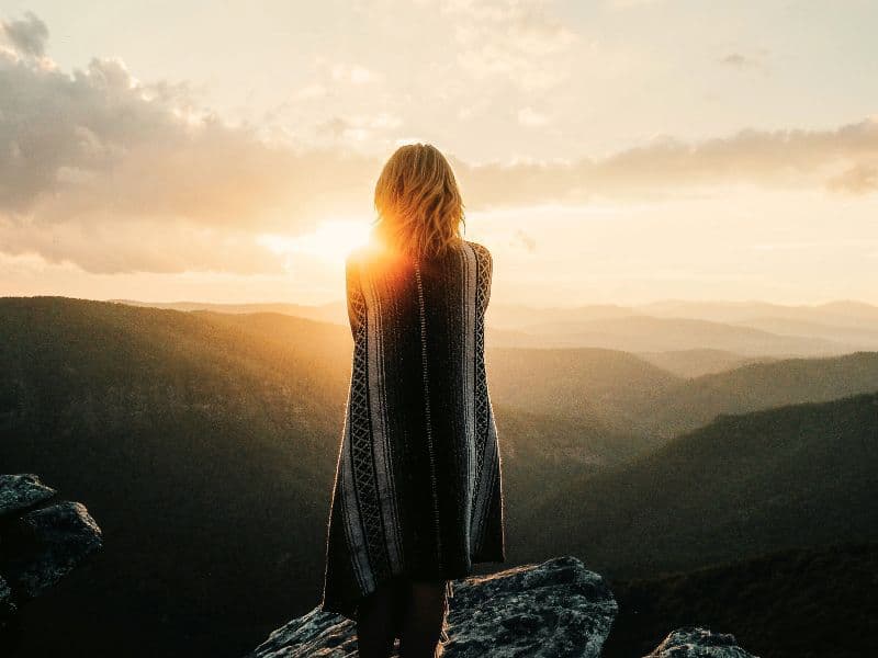 Lady standing on top of cliff staring at sunrise appearing to be reflecting.