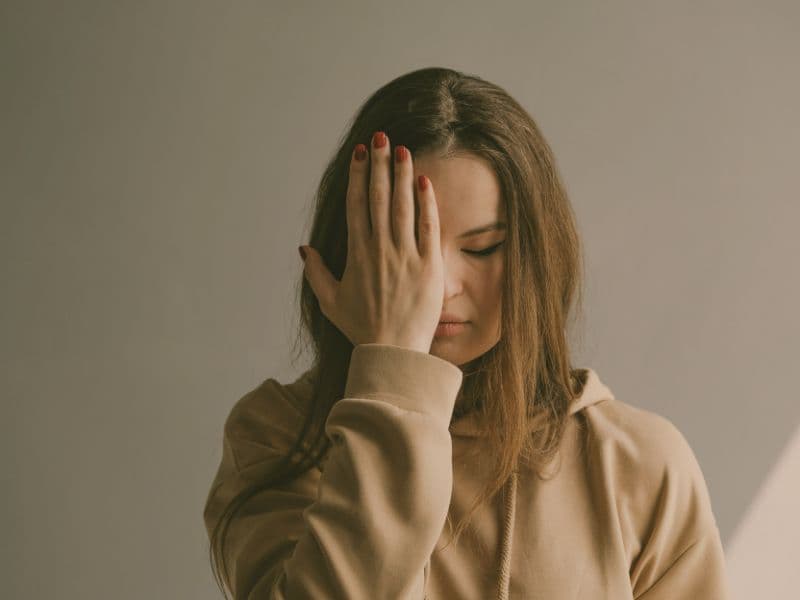 Woman looking downward with hand covering half her face, appearing overwhelmed and shameful.