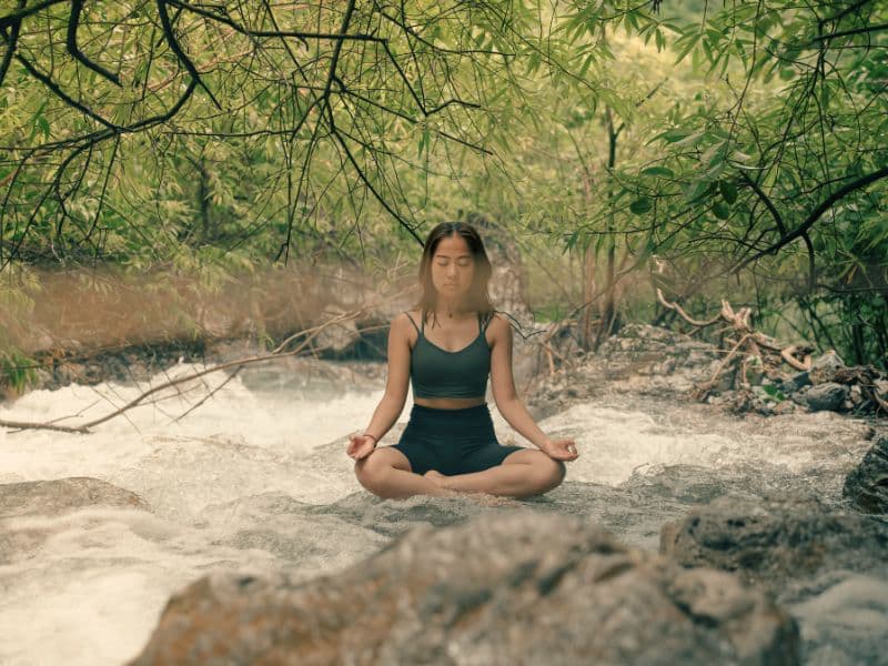 Girl meditating on rocks by the river representing healing from trauma through breathwork.