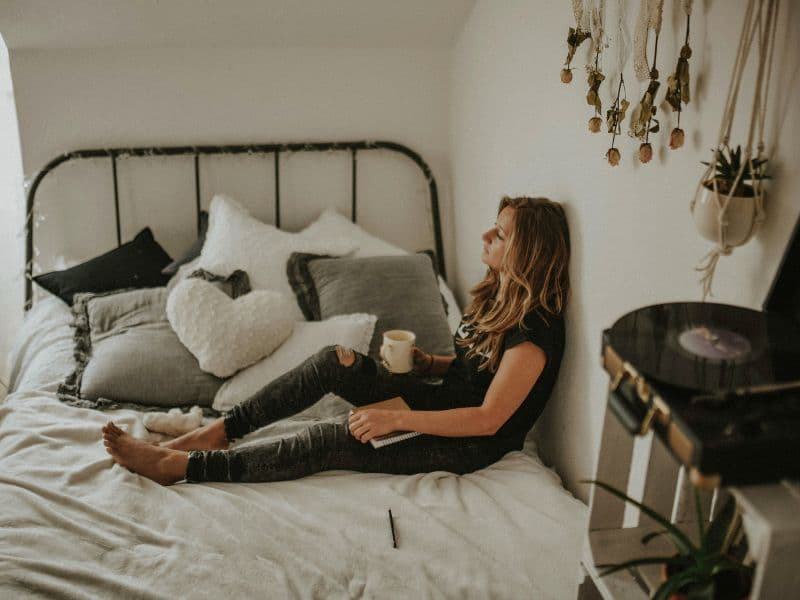 Woman sitting quietly on a bed with her back against the wall, appearing emotionally exhausted and lost in thought.