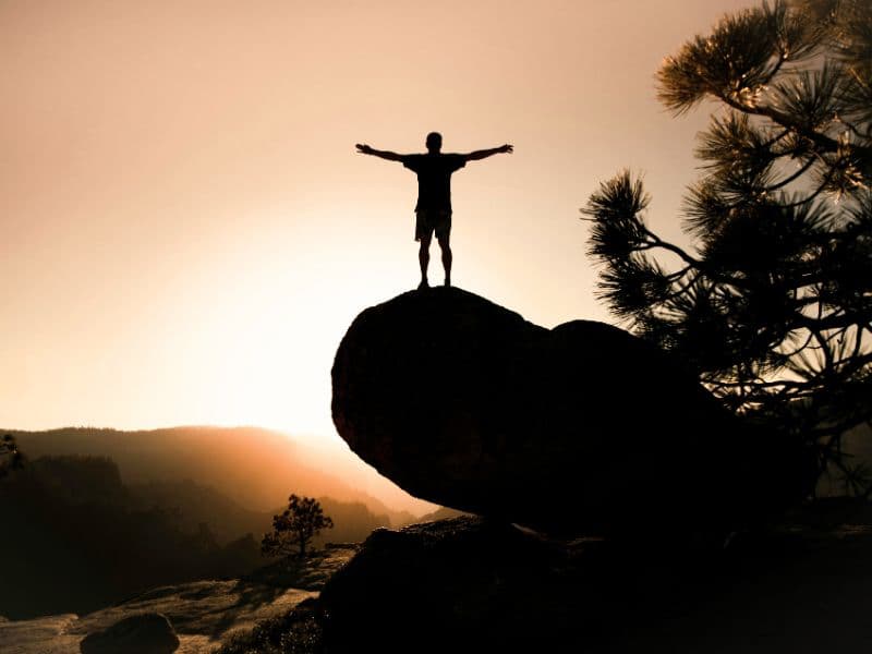Person standing on a rock overlooking a sunset with arms outstretched, representing peace and growth after healing from childhood trauma.