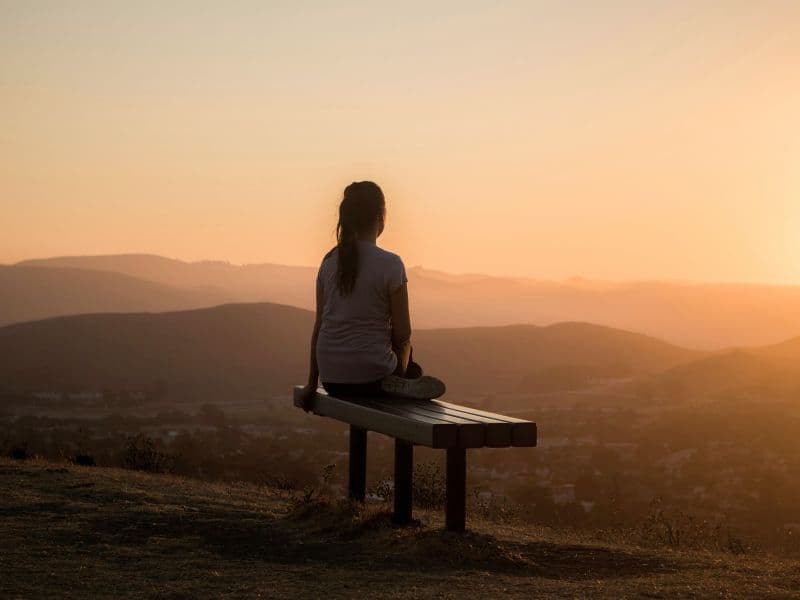 Woman sitting on a bench overlooking mountains at sunset, symbolizing hope and healing after living in survival mode.