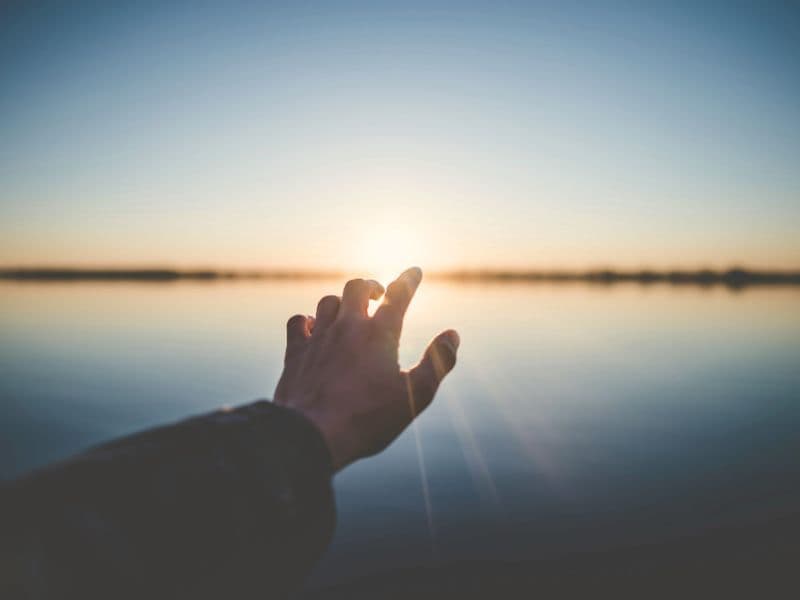 Hand reaching toward the sunrise over the ocean.